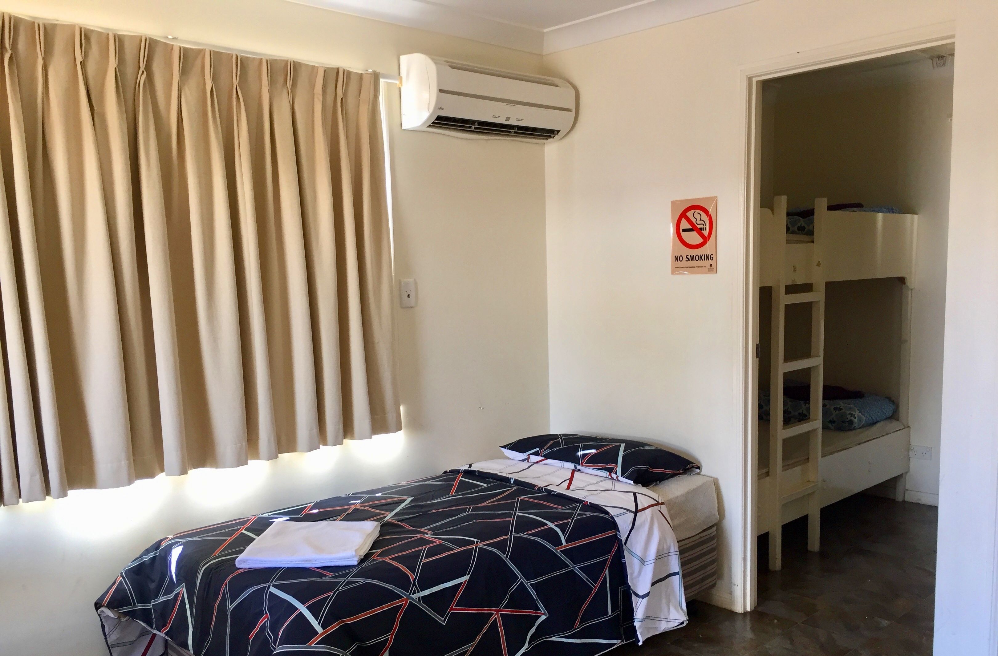 A bedroom with a bed, a window with curtains, an air conditioning unit, a bunk bed, and a 'No Smoking' sign.