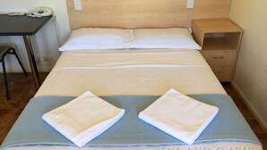 A hotel room with a wooden headboard, two white pillows, a bed with a blue and white striped bedspread, two white towels, a small wooden nightstand, a desk with a telephone, and a wall-mounted fan. - Inland Oasis Motel (Soldiers Hill)