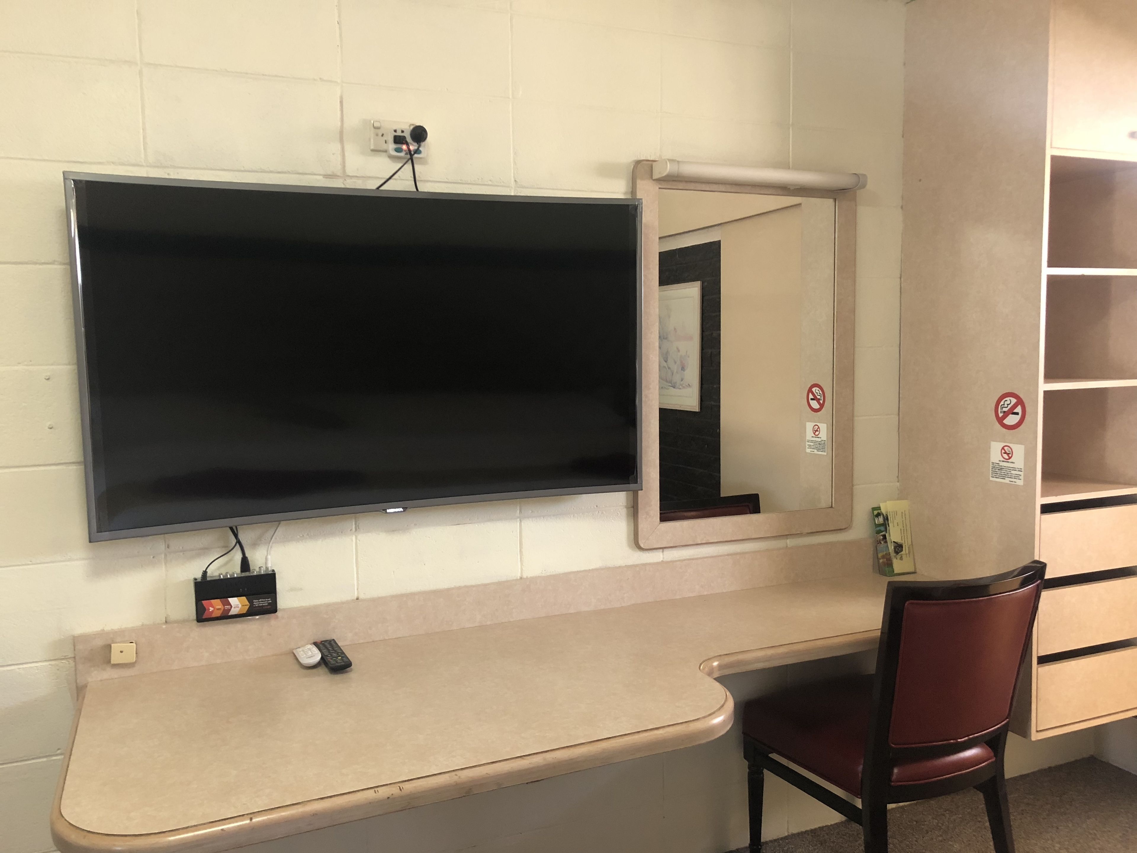 A hotel room with a flat-screen TV mounted on the wall, a desk with a chair, and a mirror.