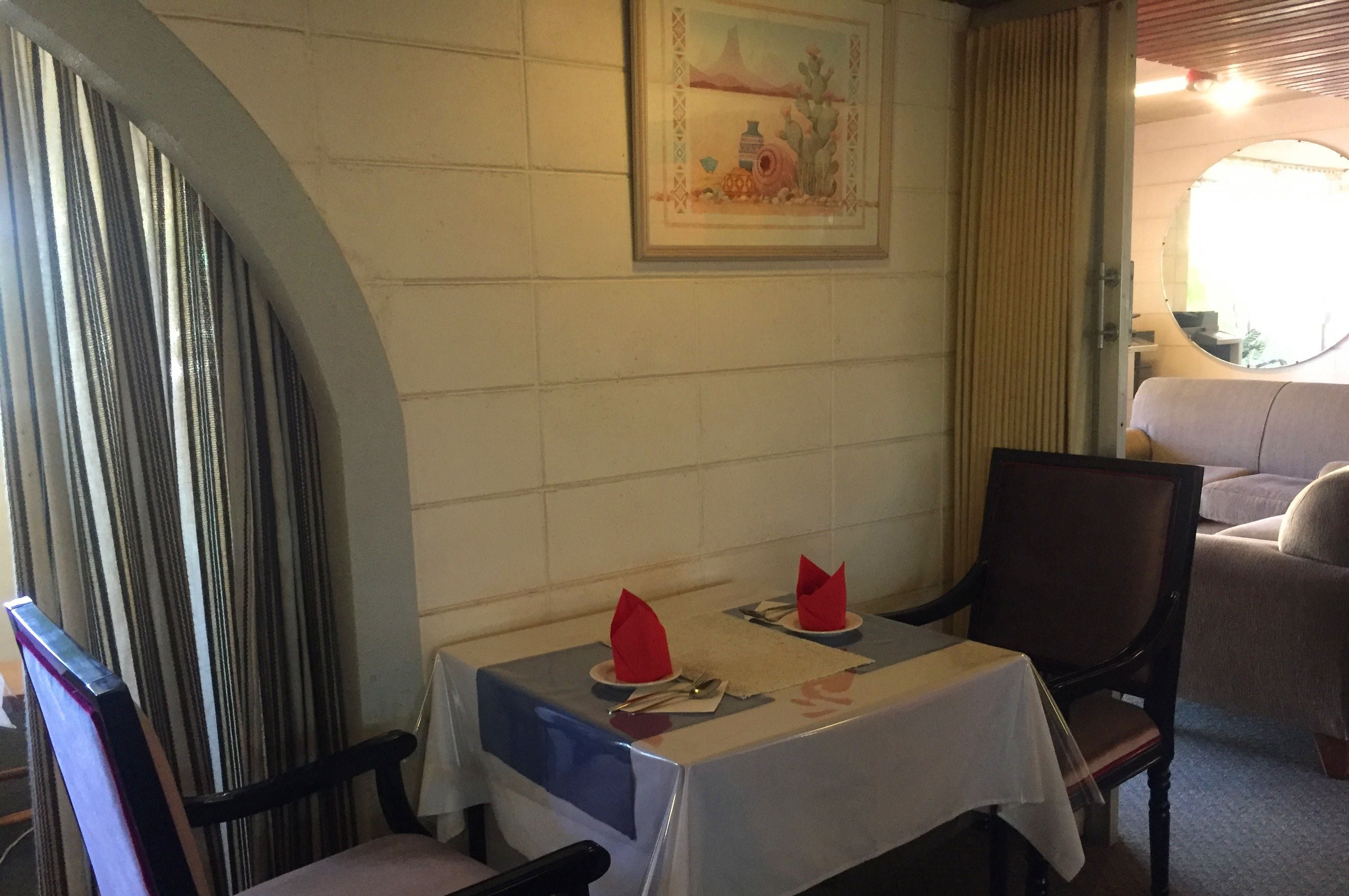 Un coin repas avec une table dressée pour deux personnes, un tableau encadré au mur et une fenêtre avec des rideaux.