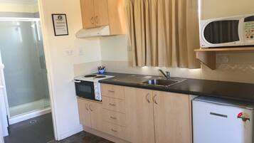 A compact kitchen with a stove, sink, and microwave. There is a small refrigerator and wooden cabinets. A shower is visible in the adjacent room.