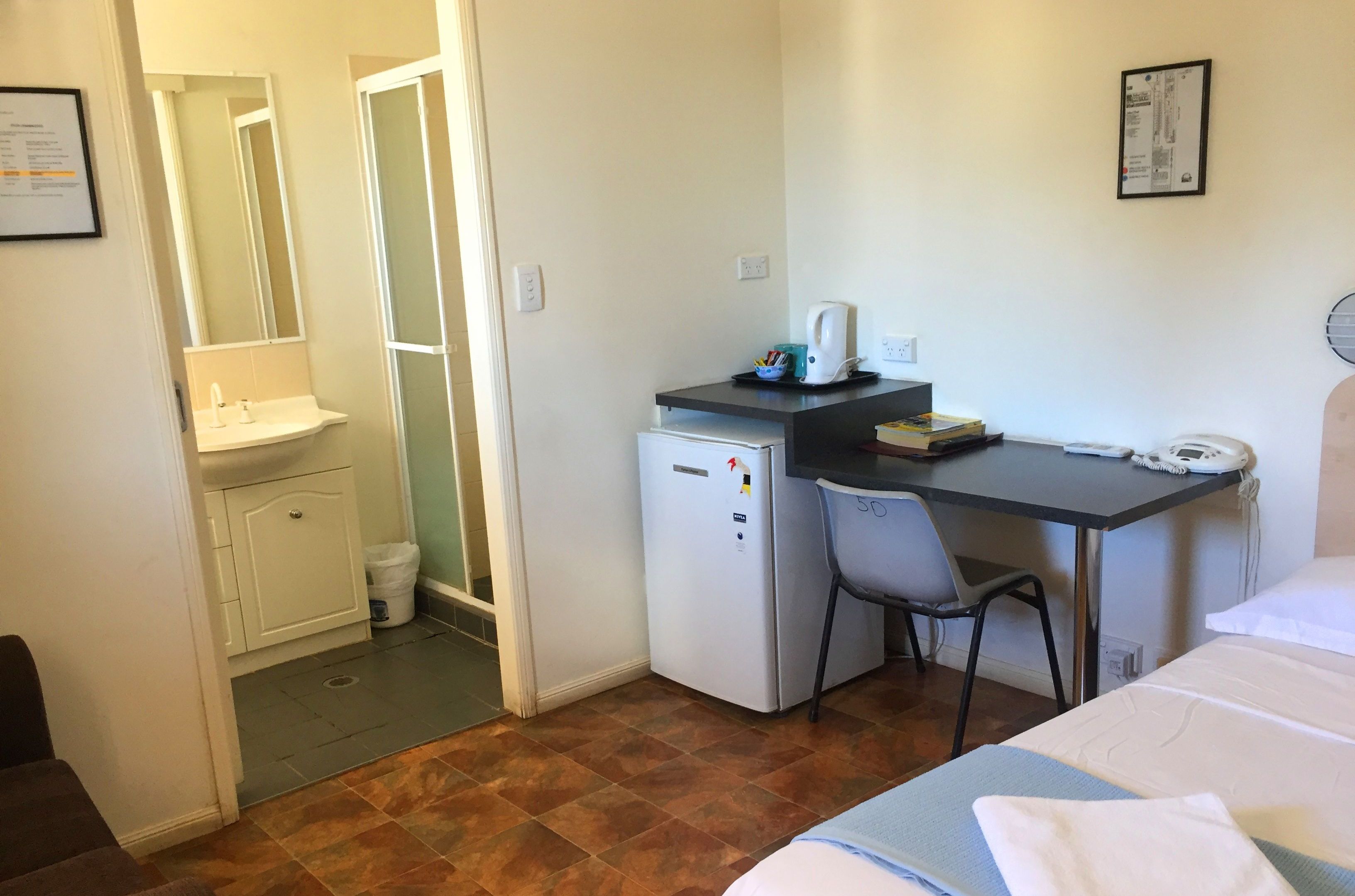 A small hotel room with a bed, a desk with a chair, a refrigerator, a sink, and a mirror.