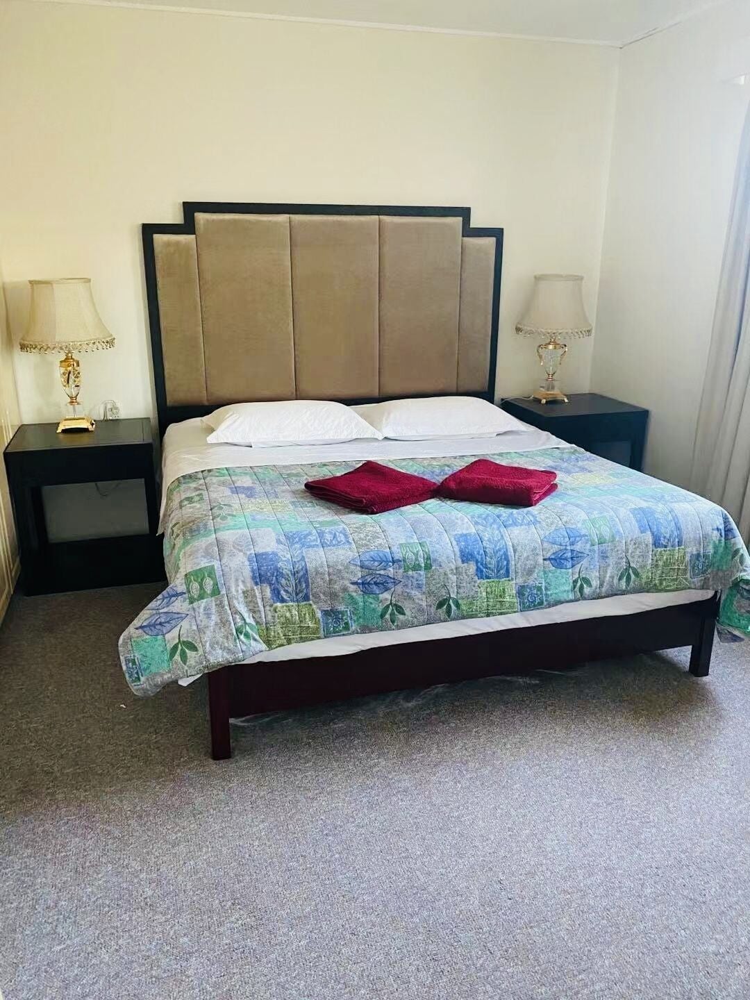 Ein ordentlich bezogenes Bett mit Blumenmusterdecke, zwei Kissen und zwei roten Handtüchern. Zwei Nachttische mit Lampen und ein dunkel gefärbtes Kopfteil sind ebenfalls vorhanden.