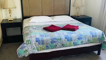 Ein ordentlich bezogenes Bett mit Blumenmusterdecke, zwei Kissen und zwei roten Handtüchern. Zwei Nachttische mit Lampen und ein dunkel gefärbtes Kopfteil sind ebenfalls vorhanden.