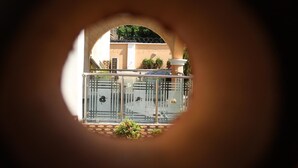 Uitzicht door een ronde opening, op een balkon met een metalen hekwerk, een plant in pot en een gebouw op de achtergrond.
