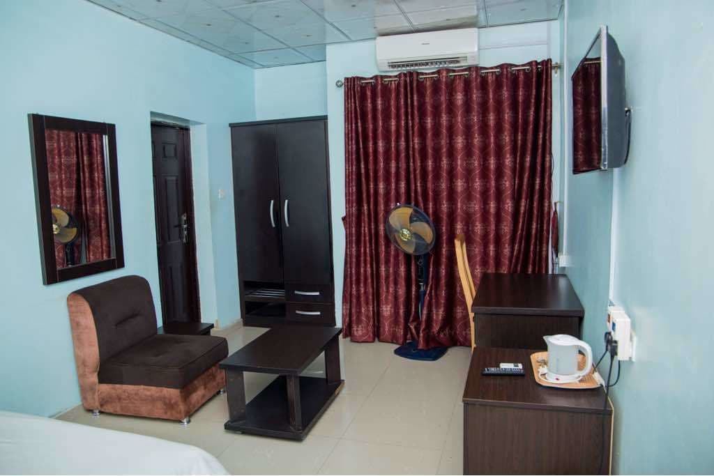 A hotel room with a bed, a sofa, a TV, a fan, and a desk with a kettle.
