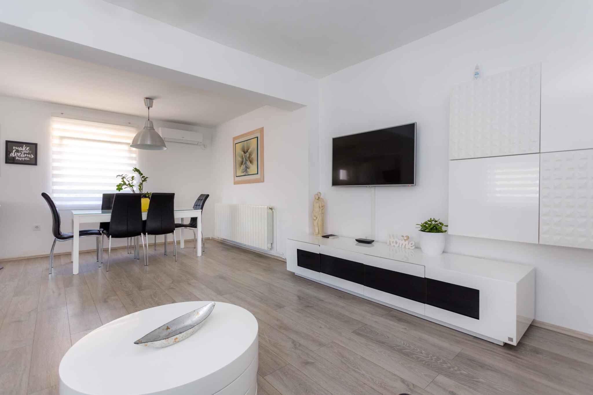 A modern living room with a dining area, white furniture, a flat-screen TV, and a radiator.
