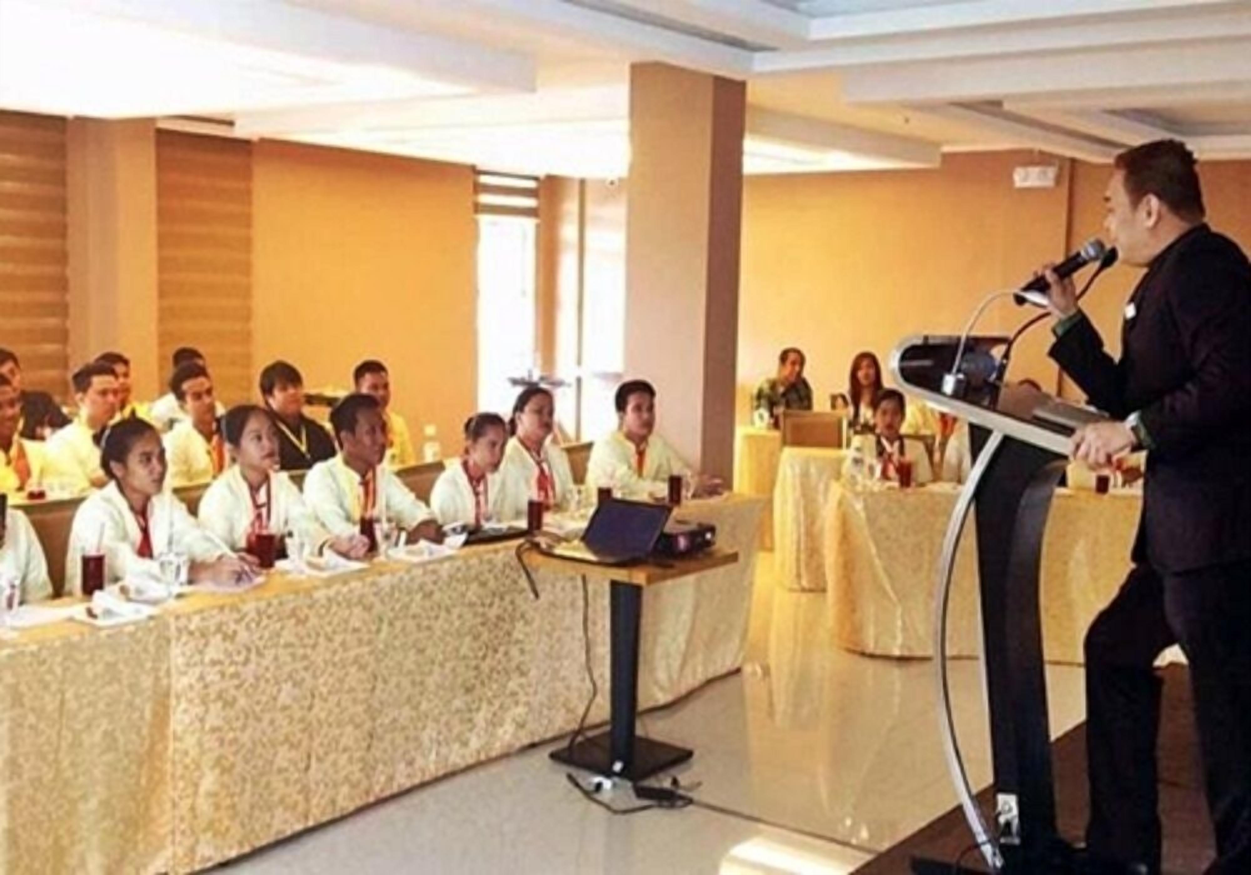 Un salón de conferencias con un orador en el atril y asistentes sentados alrededor de una mesa larga.