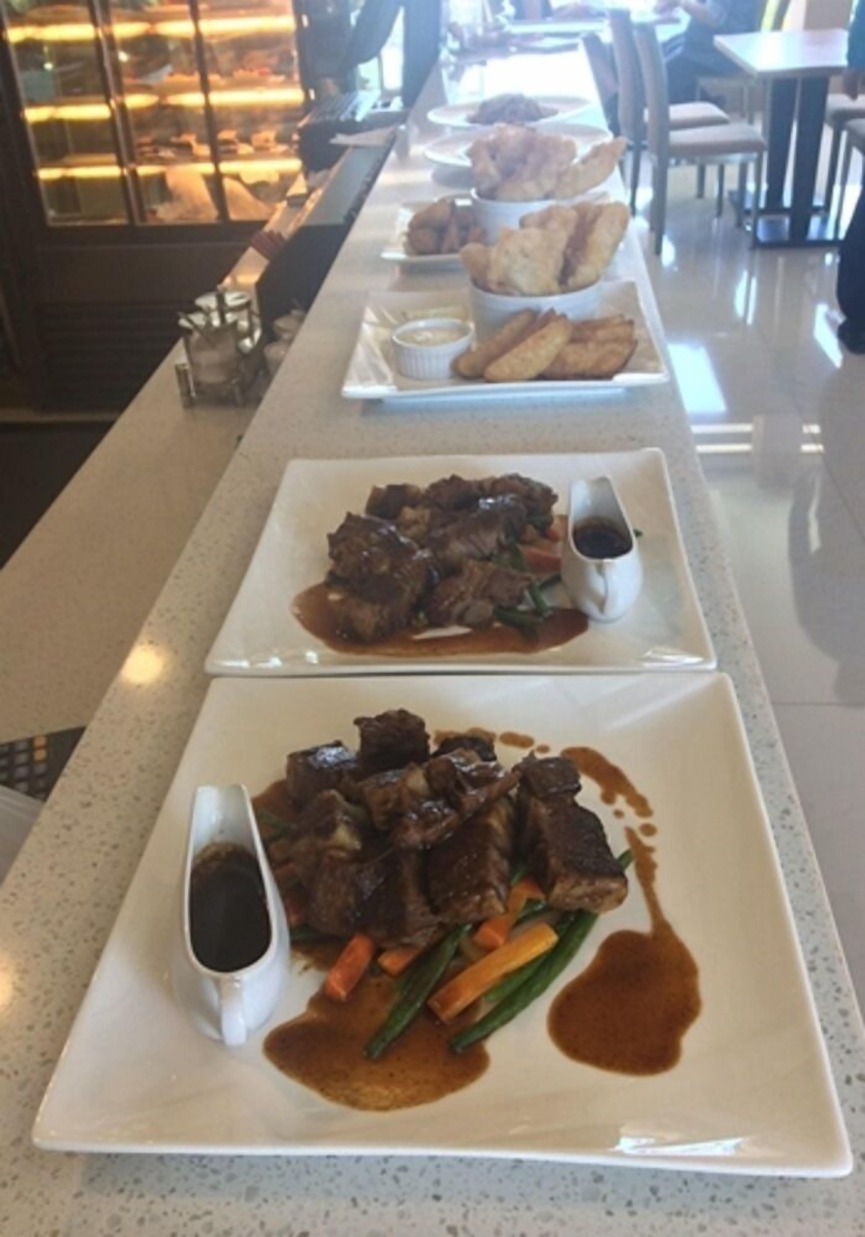 Mostrador de restaurante con varios platos, incluyendo un guiso de carne con verduras y salsa, y una bandeja de postres con pastelería.