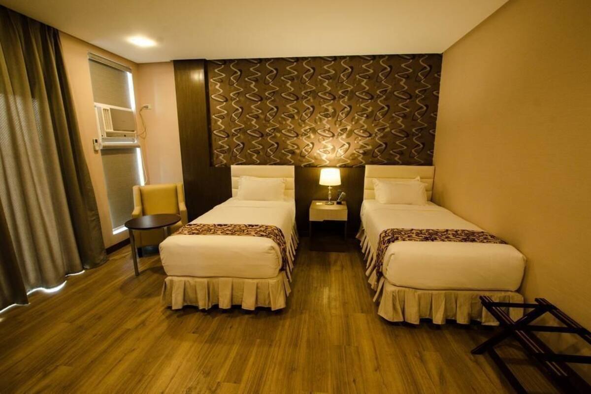 A hotel room with two beds, a small table, a chair, and a wall-mounted lamp.