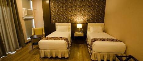 A hotel room with two beds, a small table, a chair, a nightstand with a lamp, and a patterned wall.
