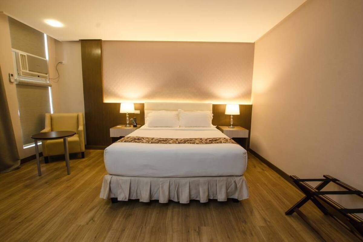 A hotel room with a large bed, two bedside lamps, a small round table, a chair, and a wooden floor.