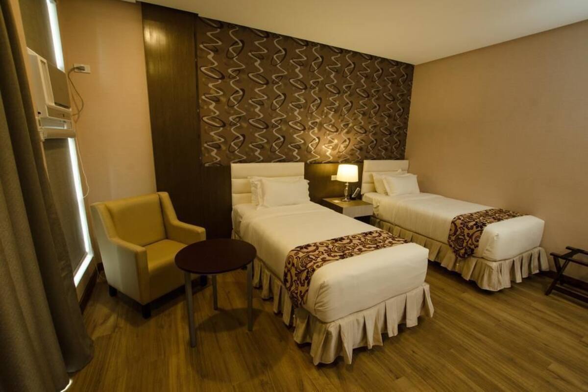 A hotel room with two beds, a small armchair, a bedside table with a lamp, and a patterned wall.