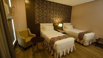 Habitación de hotel con dos camas, un sillón pequeño, una mesita de noche con lámpara y un papel tapiz estampado.