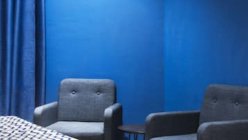 Habitación de hotel con una pared azul, dos sillones grises, una mesita y una cama con colcha estampada.