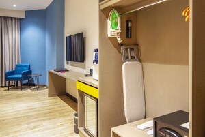 Habitación de hotel con pared azul, suelo de madera, una silla azul, escritorio con computadora, televisor, secador de pelo y espejo.