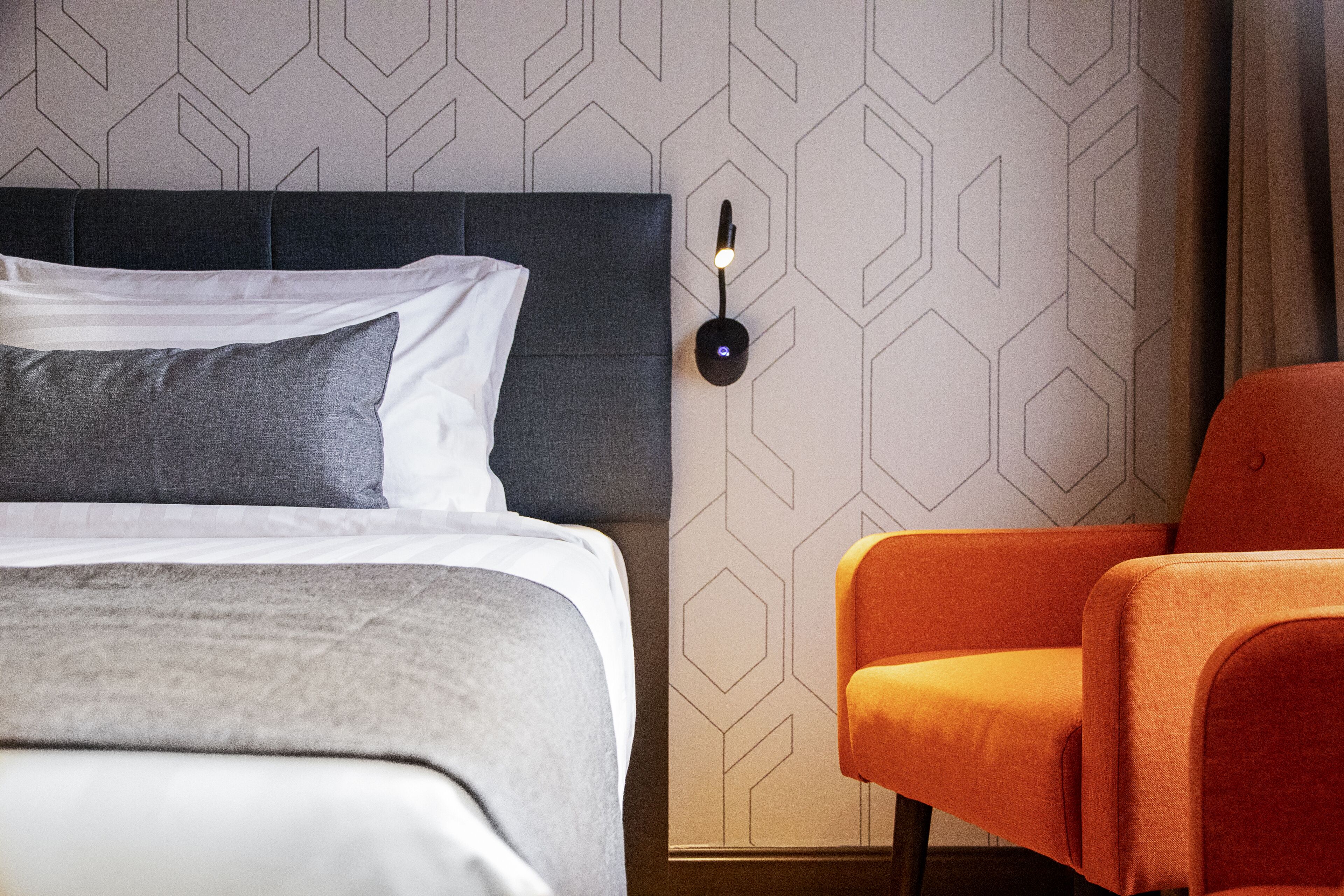 A modern hotel room with a neatly made bed, a stylish orange armchair, and a geometric-patterned wall.