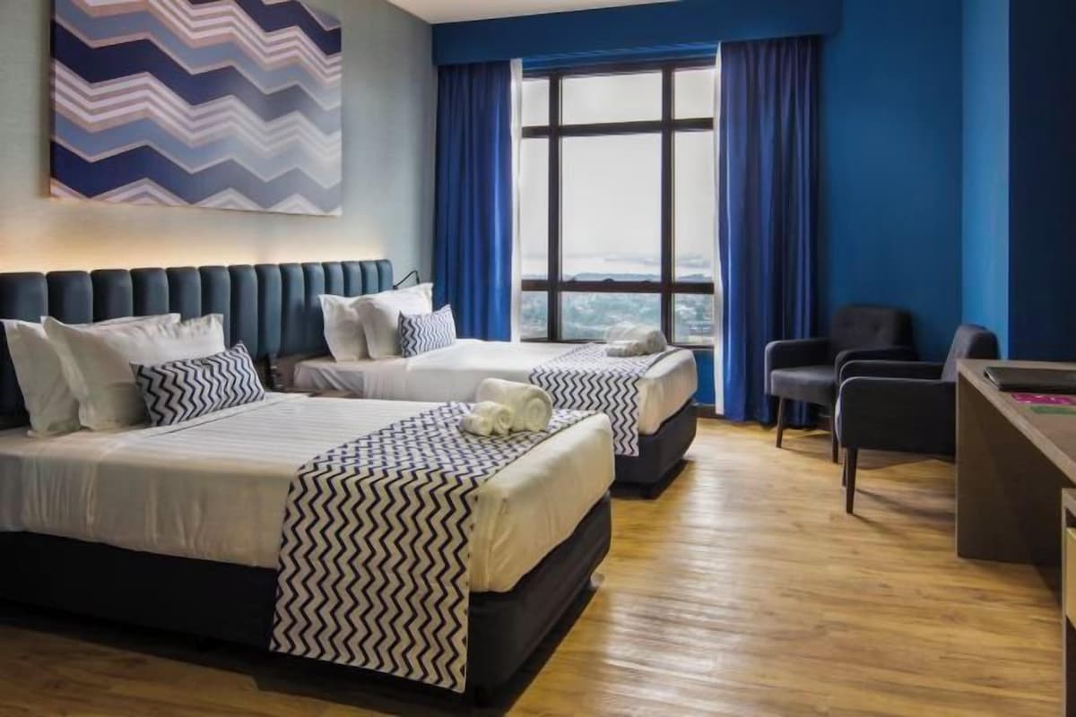 A hotel room with two beds, a desk, and a view through large windows.