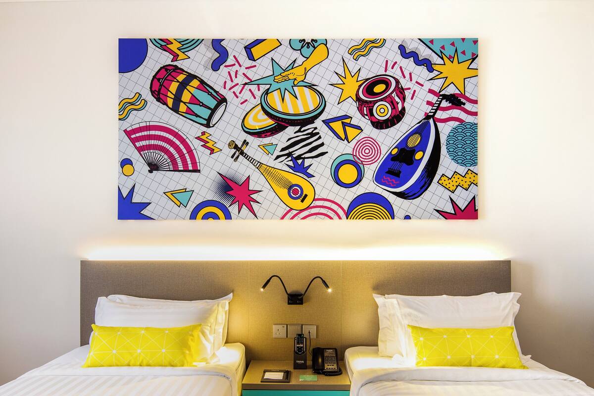A hotel room with two beds, each with white linens and a yellow pillow. A colorful abstract painting decorates the wall above the beds.