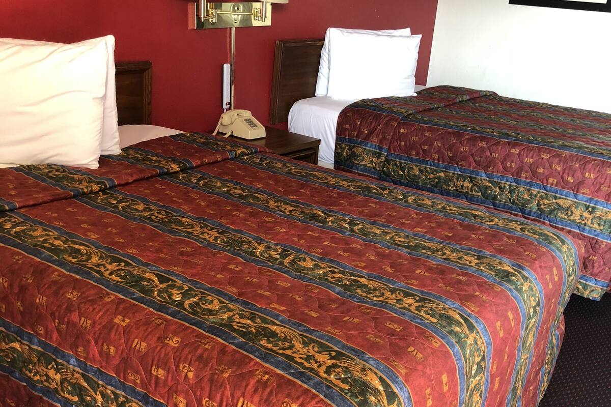 A hotel room with two beds, each with a red and gold patterned bedspread, a red wall, a lamp, and a framed picture on the wall.