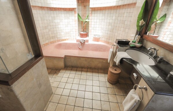 Un baño con bañera, lavamanos y espejo. Hay plantas en macetas y una cesta en el suelo.