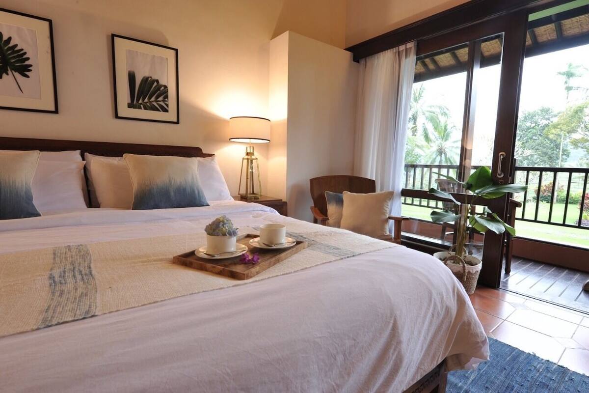 A hotel room with a large bed, a nightstand with a lamp, a chair, a balcony with a view of palm trees, and a potted plant.