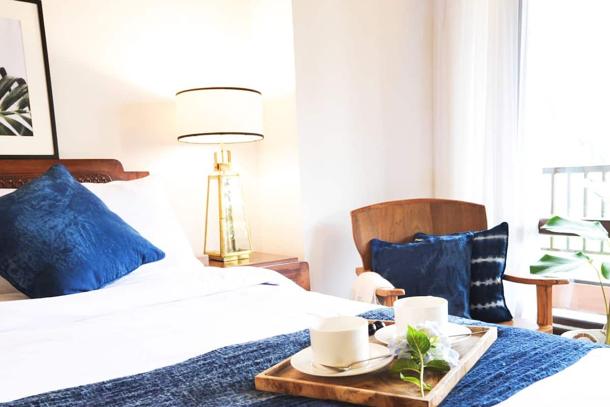 A hotel room with a bed, a nightstand with a lamp, a chair, and a tray with a cup and flowers.