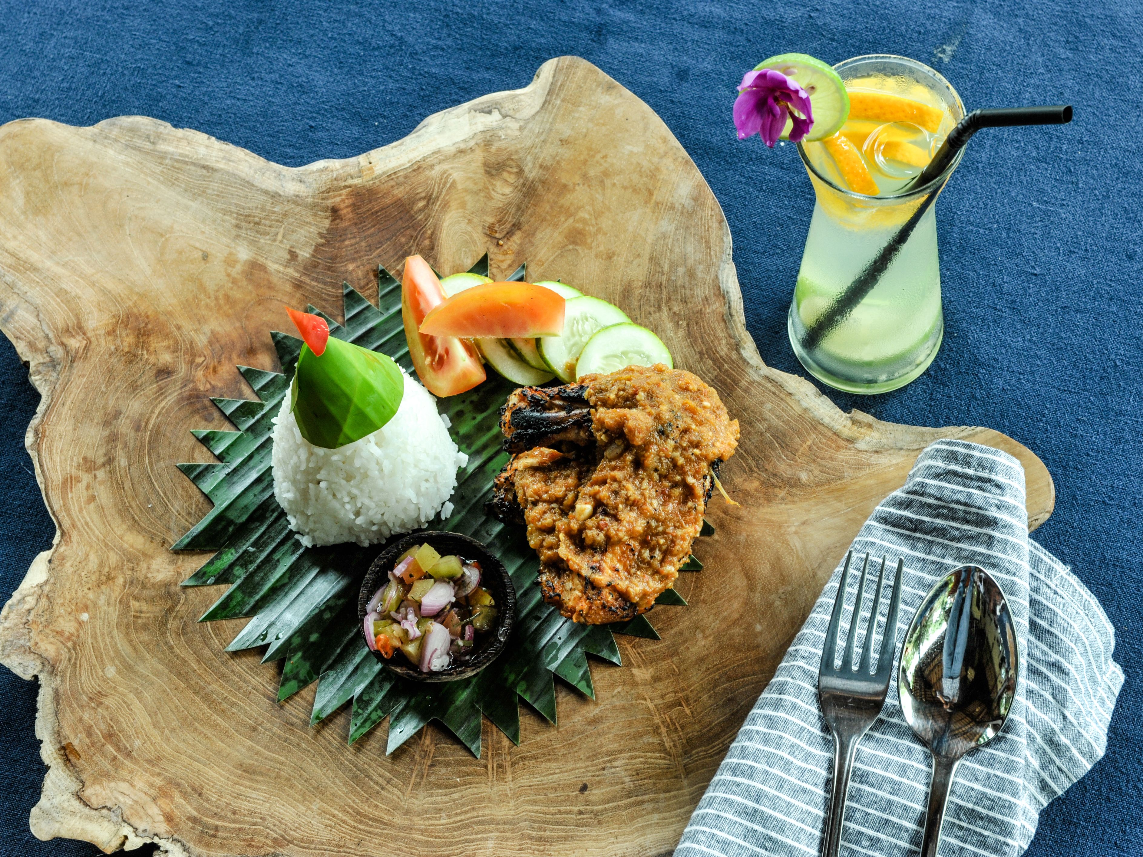 Una tabla de madera con arroz blanco, pollo a la parrilla y un recipiente pequeño con sambal.