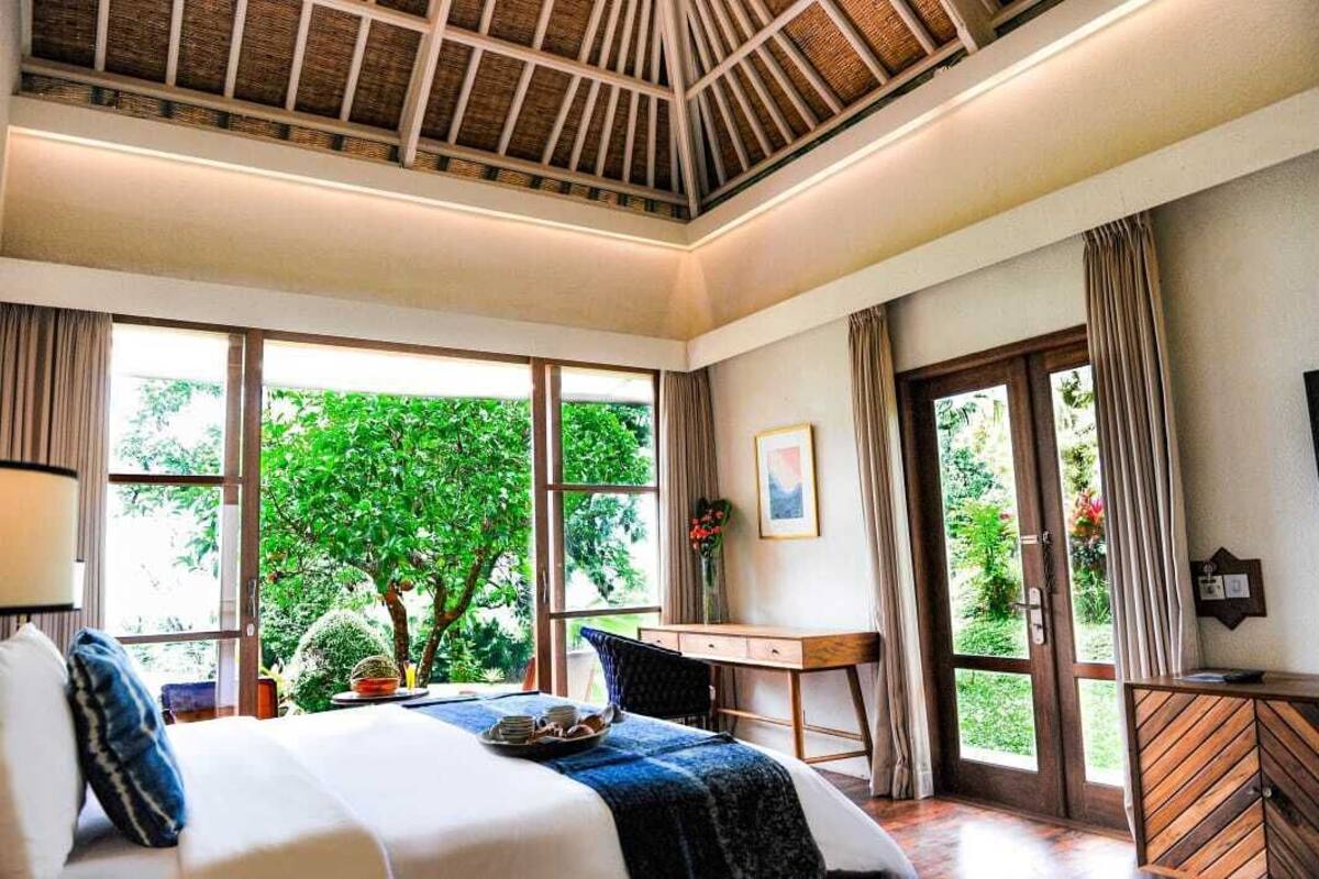 A bedroom with a large bed, a desk, and a view of greenery through the windows.