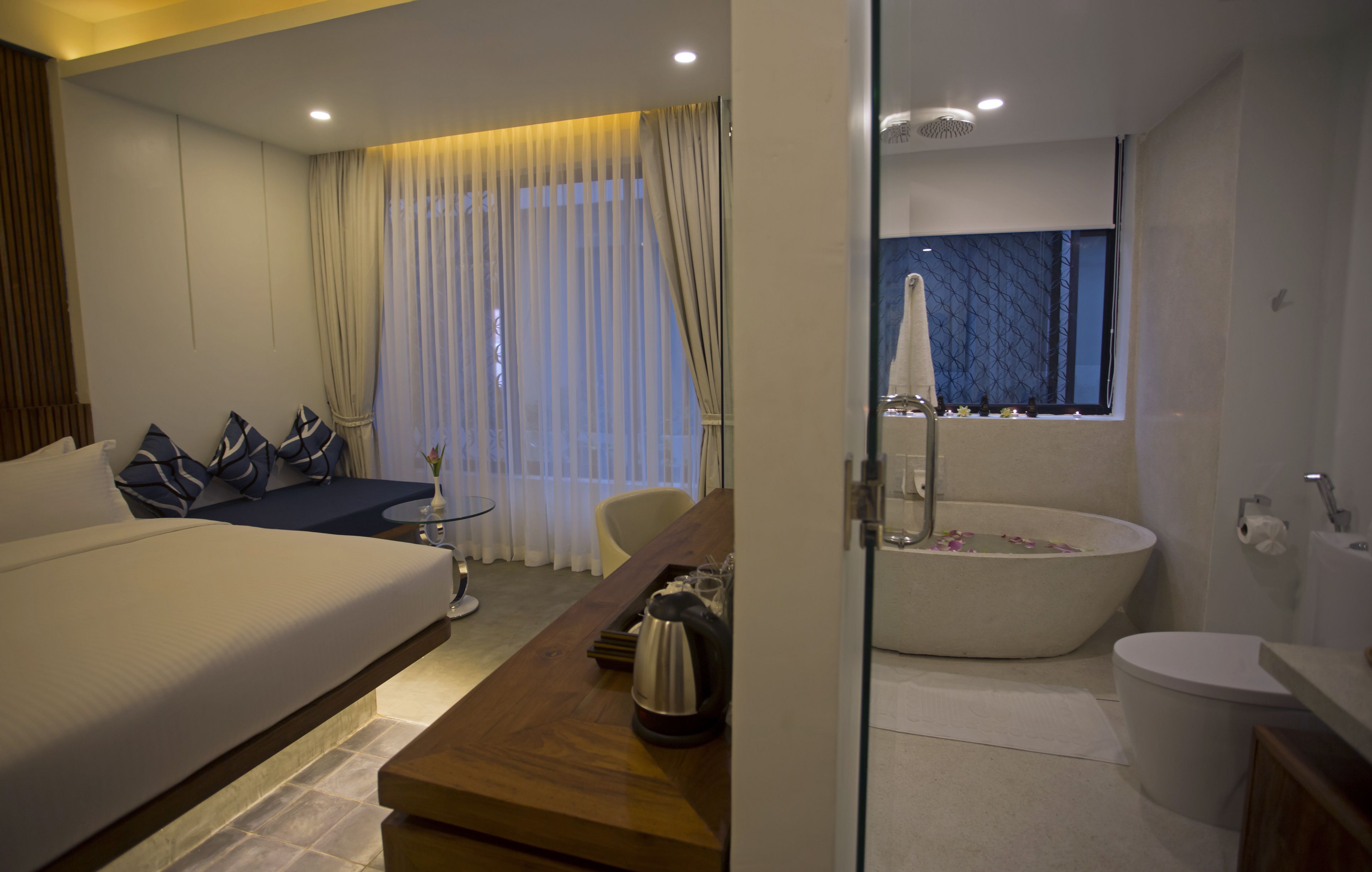A modern hotel room with a large bed, a wooden desk, a sofa with patterned cushions, a glass-enclosed bathroom with a bathtub and toilet, and a large window with curtains.