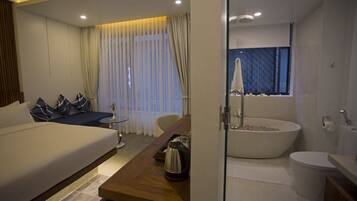 A modern hotel room with a large bed, a wooden desk, a sofa with patterned cushions, a glass-enclosed bathroom with a bathtub and toilet, and a large window with curtains.
