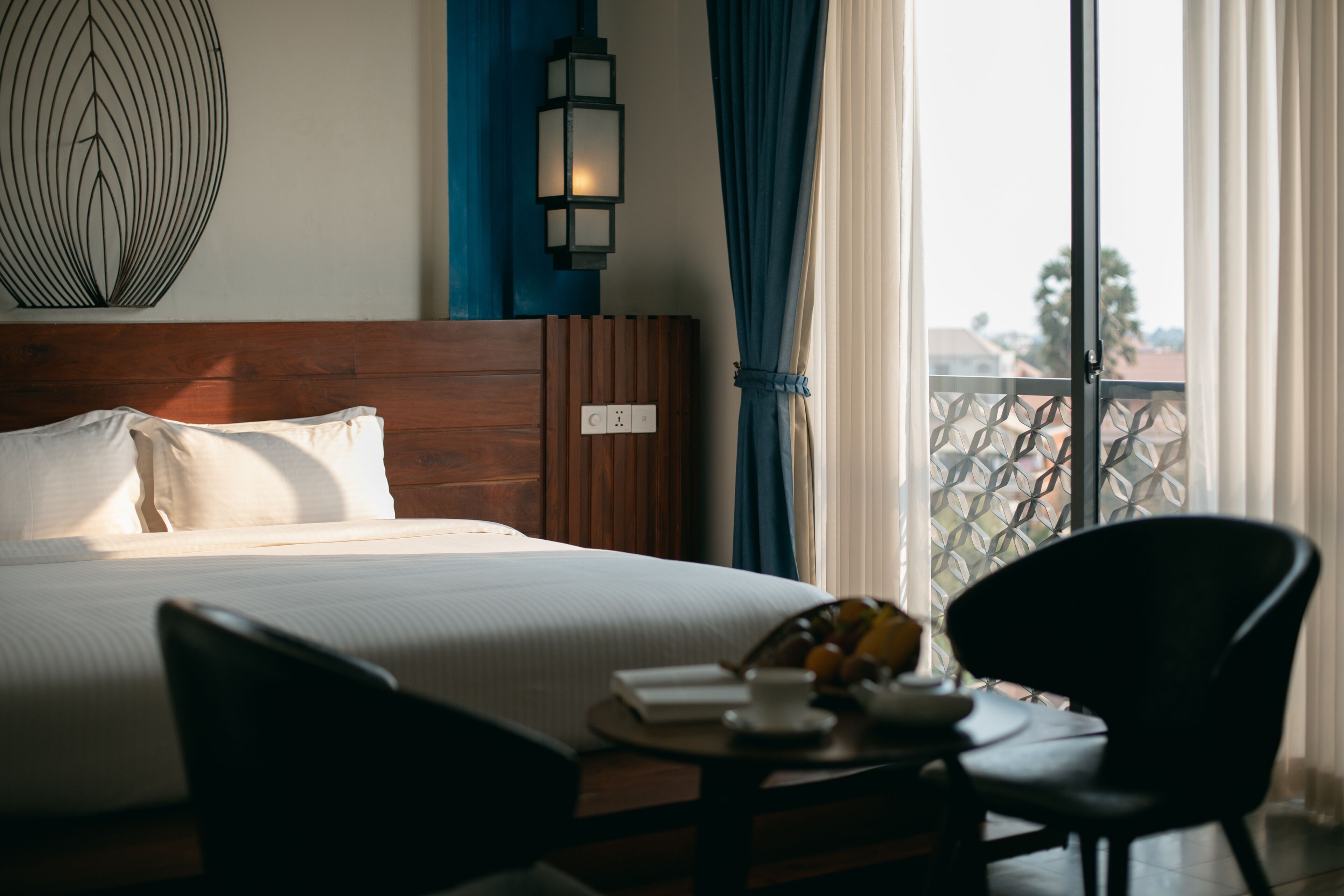 Hotelkamer met houten hoofdeinde, bed met witte lakens, een klein tafeltje met een fruitschaal, een stoel en uitzicht door een raam met balkon.