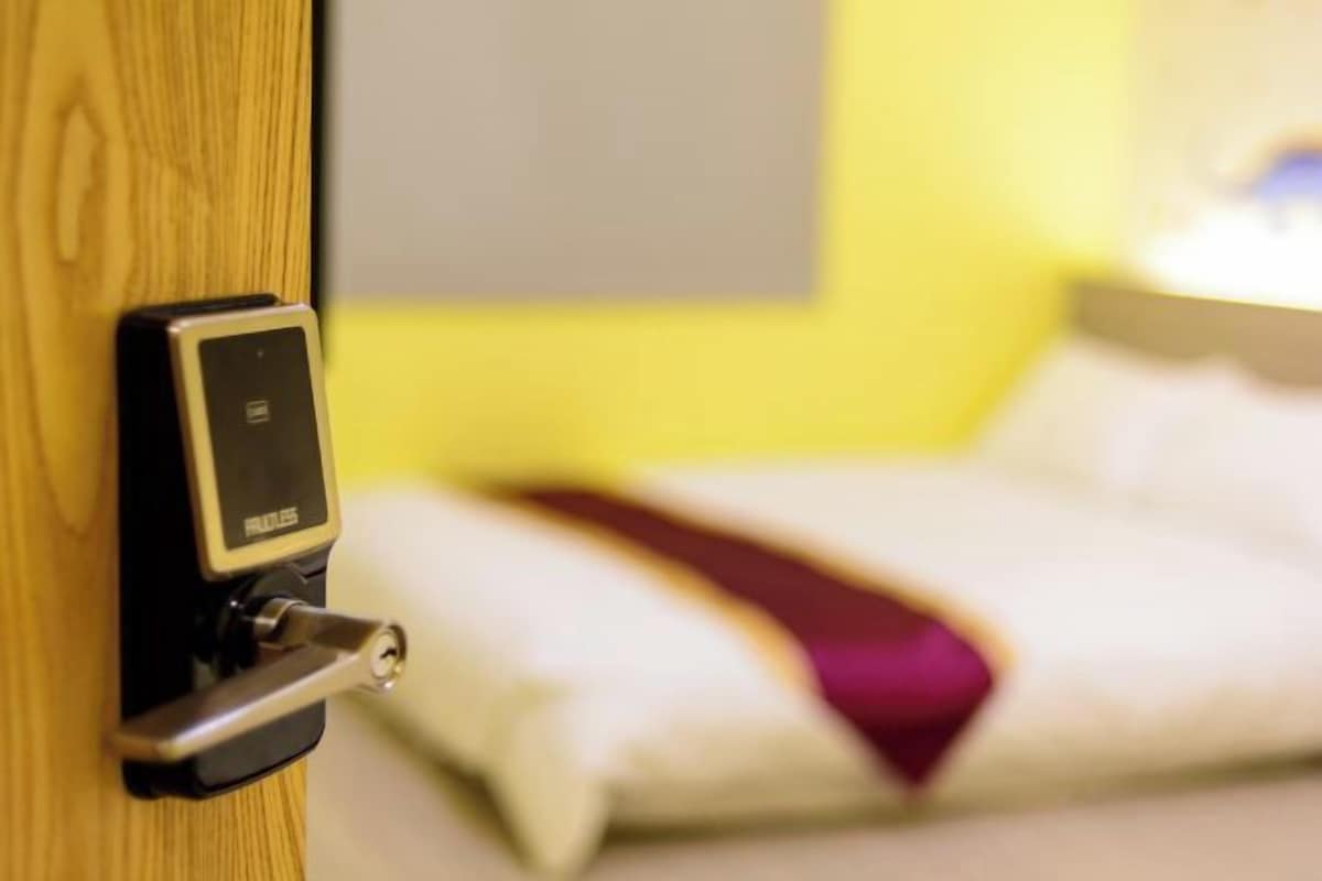 A hotel room with a wooden door, a bed with white bedding and a red throw blanket, and a control panel on the door.