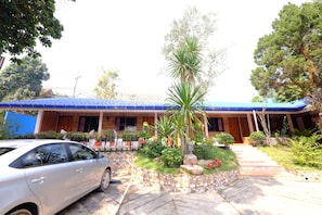 Un edificio de dos pisos con techo azul, un coche estacionado adelante y un jardín con diversas plantas y una palmera.