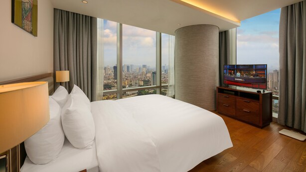 A hotel room with a large bed, a TV, and a view of a cityscape through large windows. - Lotte The Residence (Hanoi)