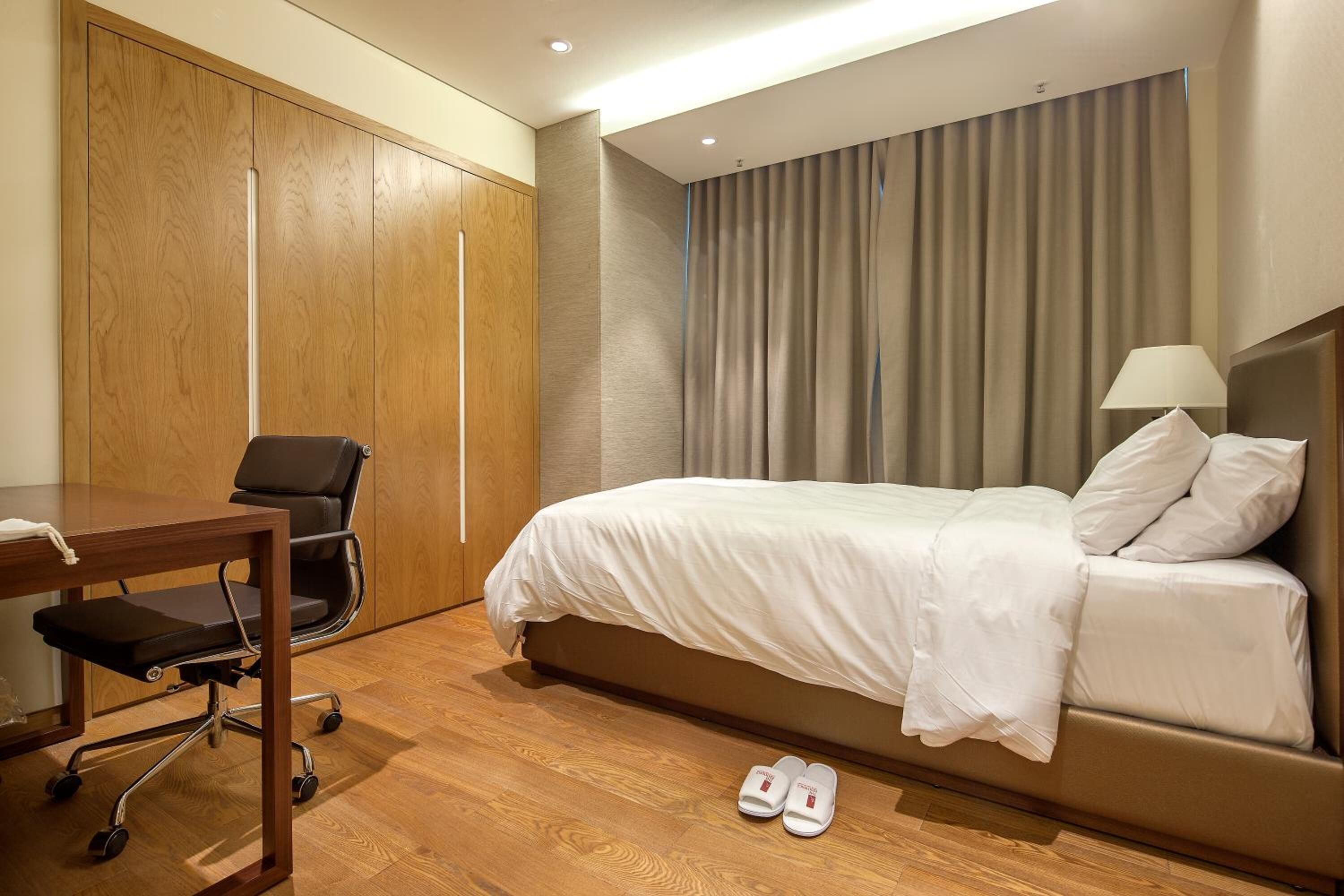 A hotel room with a bed, a desk with a chair, and a pair of slippers on the floor.