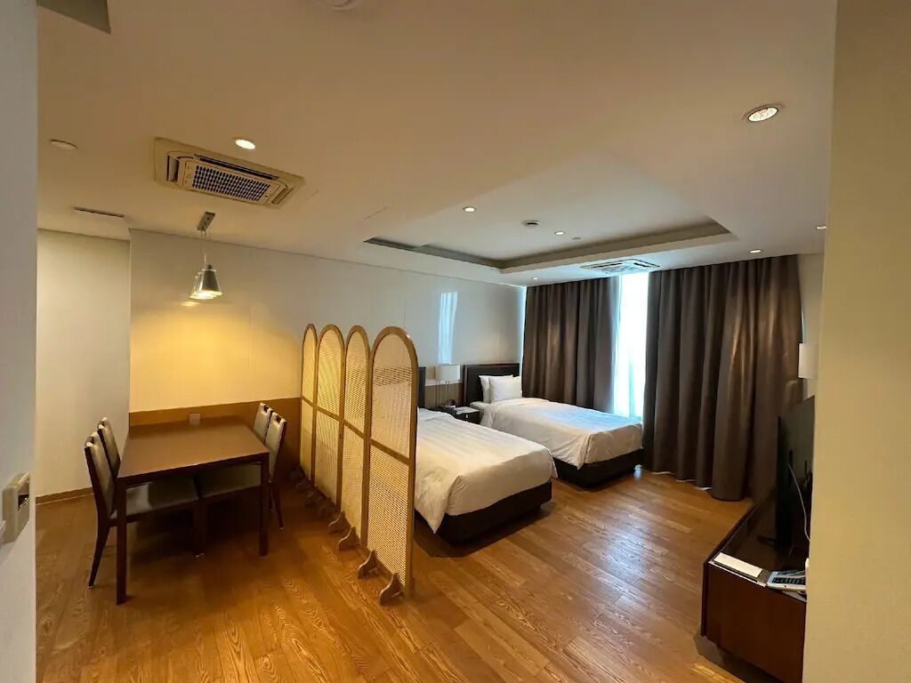 A hotel room with a wooden floor, a dining table with chairs, two single beds, a TV, and a window with curtains.