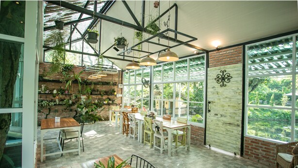 A spacious indoor area with a wooden table and chairs, a brick wall with hanging plants, and large windows offering a view of greenery outside. - Ginger Bread House - Pet Lover Resort (Nakhon Nayok)