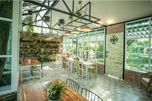 Een ruime binnenruimte met een houten tafel en stoelen, een bakstenen muur met hangplanten, en grote ramen met uitzicht op groen buiten.