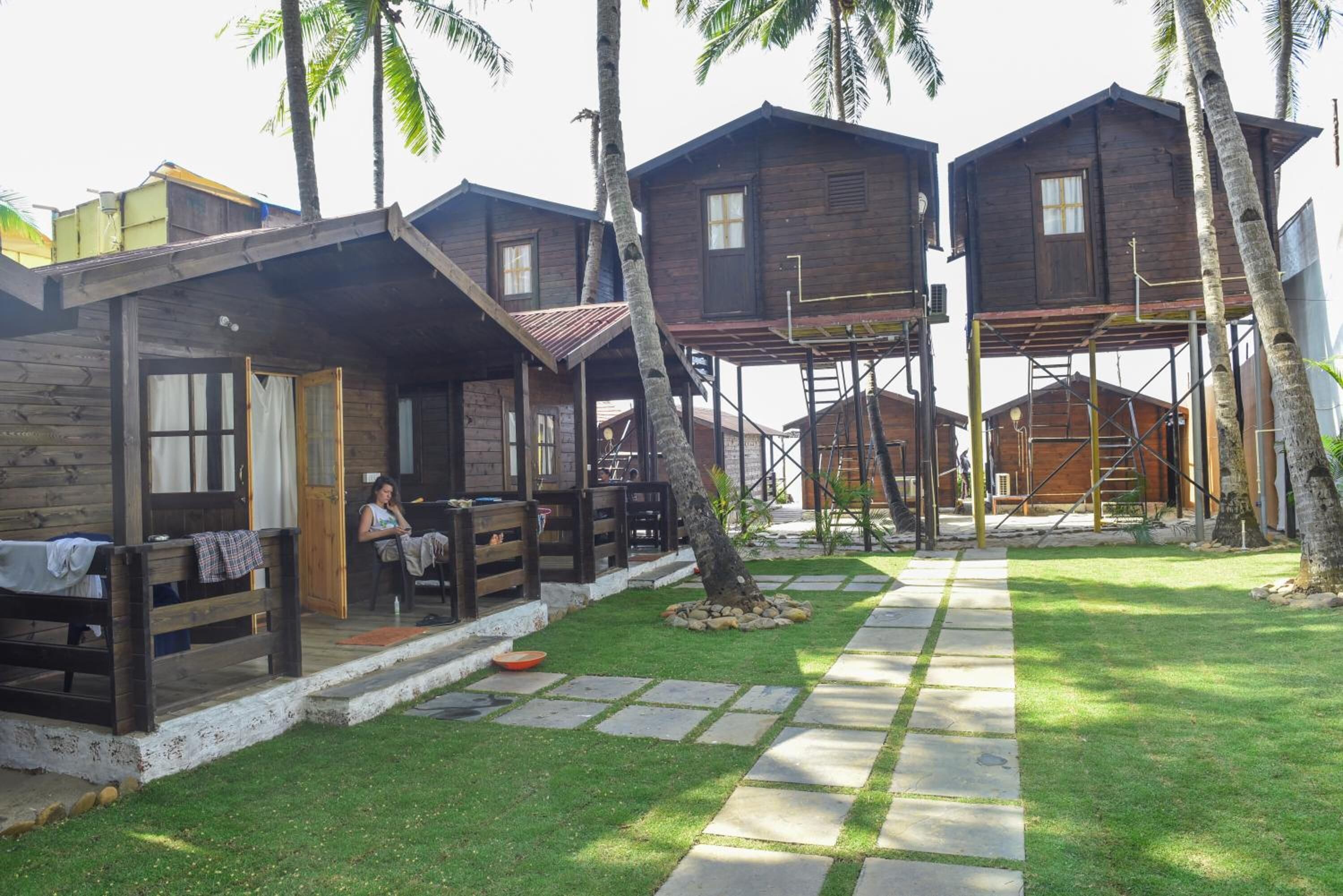 A wooden cabin-style accommodation with a covered outdoor seating area, a paved pathway, and palm trees.
