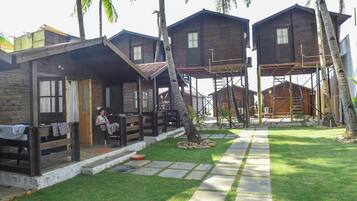 A wooden cabin-style accommodation with a covered outdoor seating area, a paved pathway, and palm trees.