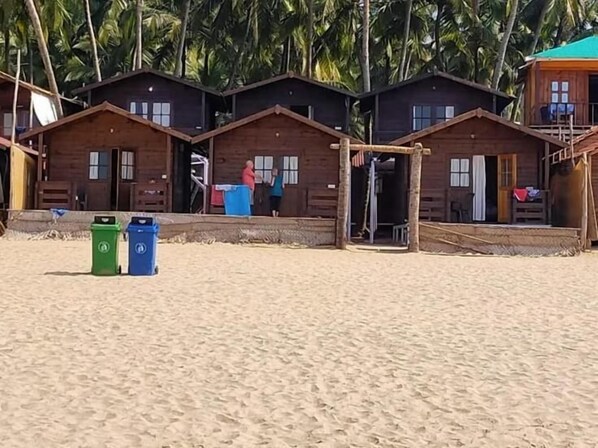 A row of wooden beach huts with a sandy beach and palm trees in the background.