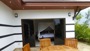 A wooden outdoor seating area with a round table and chairs, a bed with a canopy, and a sliding glass door leading to a bedroom. - Tropicana Beach Villa at  VIP Resort (Rayong)