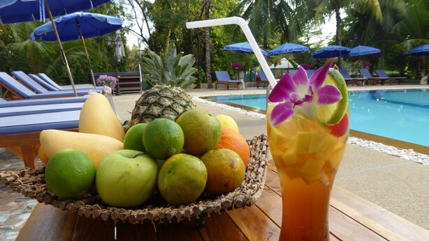 A tropical setting with a pool, lounge chairs, and a fruit bowl with a drink. - Tropicana Beach Villa at  VIP Resort (Rayong)