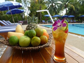 Un cadre tropical avec une piscine, des chaises longues et un panier de fruits accompagné d’une boisson.