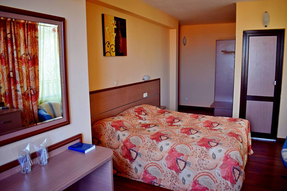 Habitación de hotel con una cama, una mesita de noche con vasos, un espejo, un cuadro en la pared y una puerta.