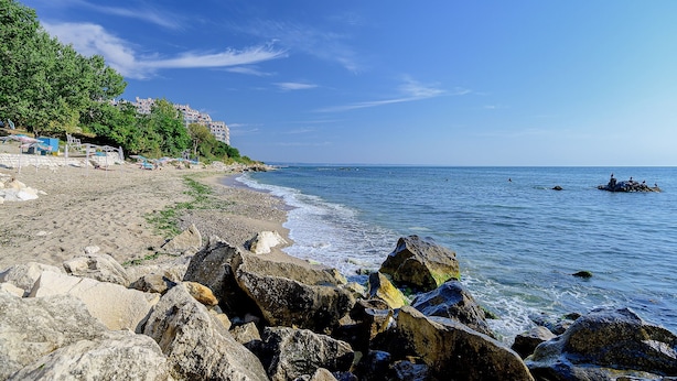 A rocky beach with a clear view of the sea and a building in the background. - Hotel Royal Beach (Varna)