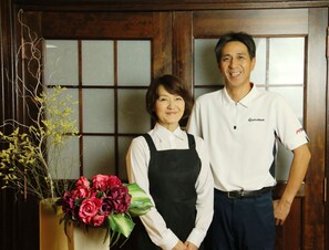 A man and a woman standing in front of a wooden door with a floral arrangement. - Hakuba Tsugaike Sunrise TANNE (Otari)