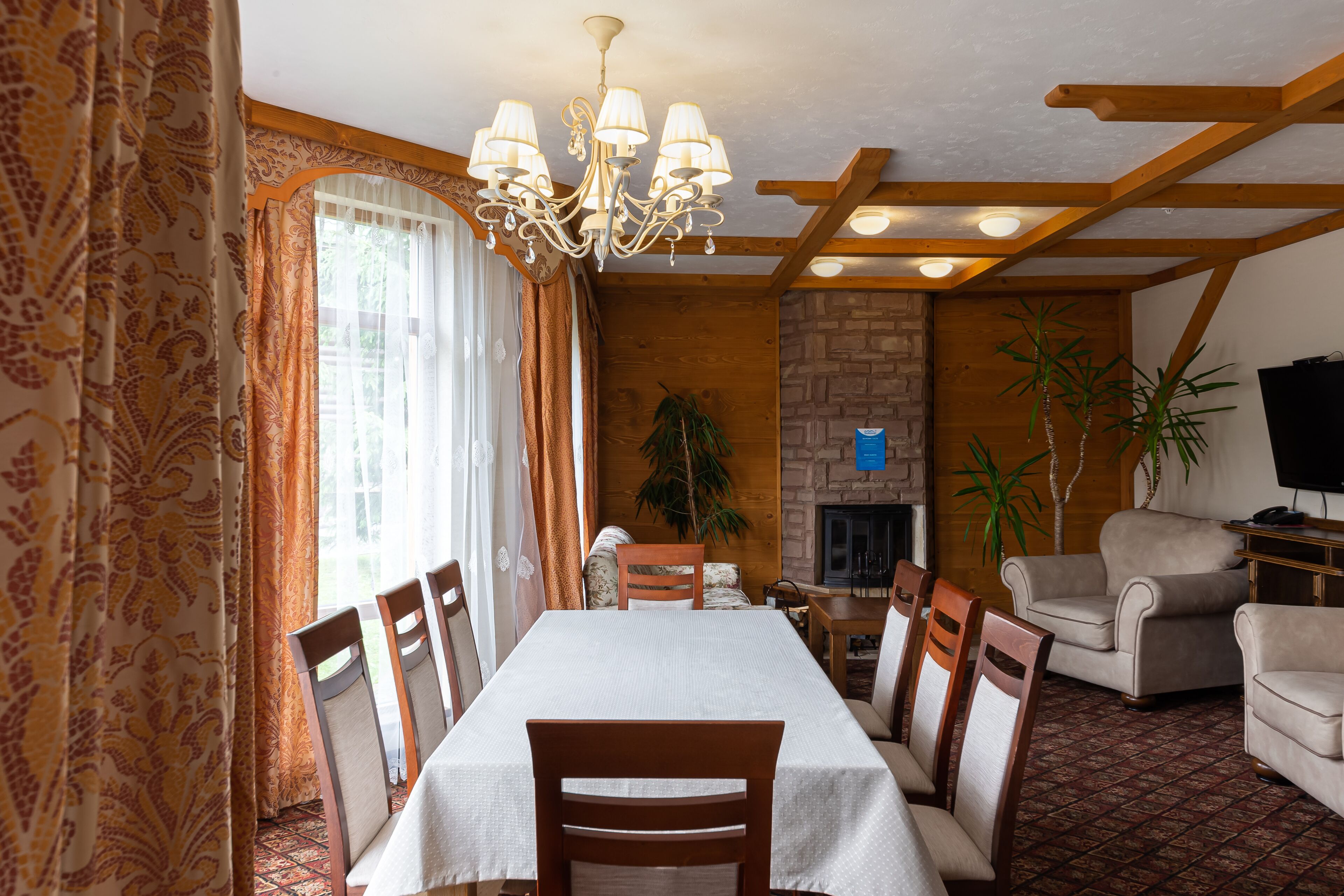 Un comedor con una mesa dispuesta para seis personas, sillas de madera, una araña, una chimenea y un televisor en una sala de estar con pared de piedra y vigas de madera.