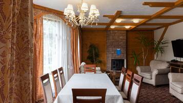 Un comedor con una mesa dispuesta para seis personas, sillas de madera, una araña, una chimenea y un televisor en una sala de estar con pared de piedra y vigas de madera.